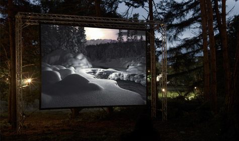 Dusk image, green and brown forest area, metal frame suspending a projector screen showing a black and white image of a snowy forest, stream and waters edge of crisp white snow, ground spotlights shining behind the metal frame, clear sky