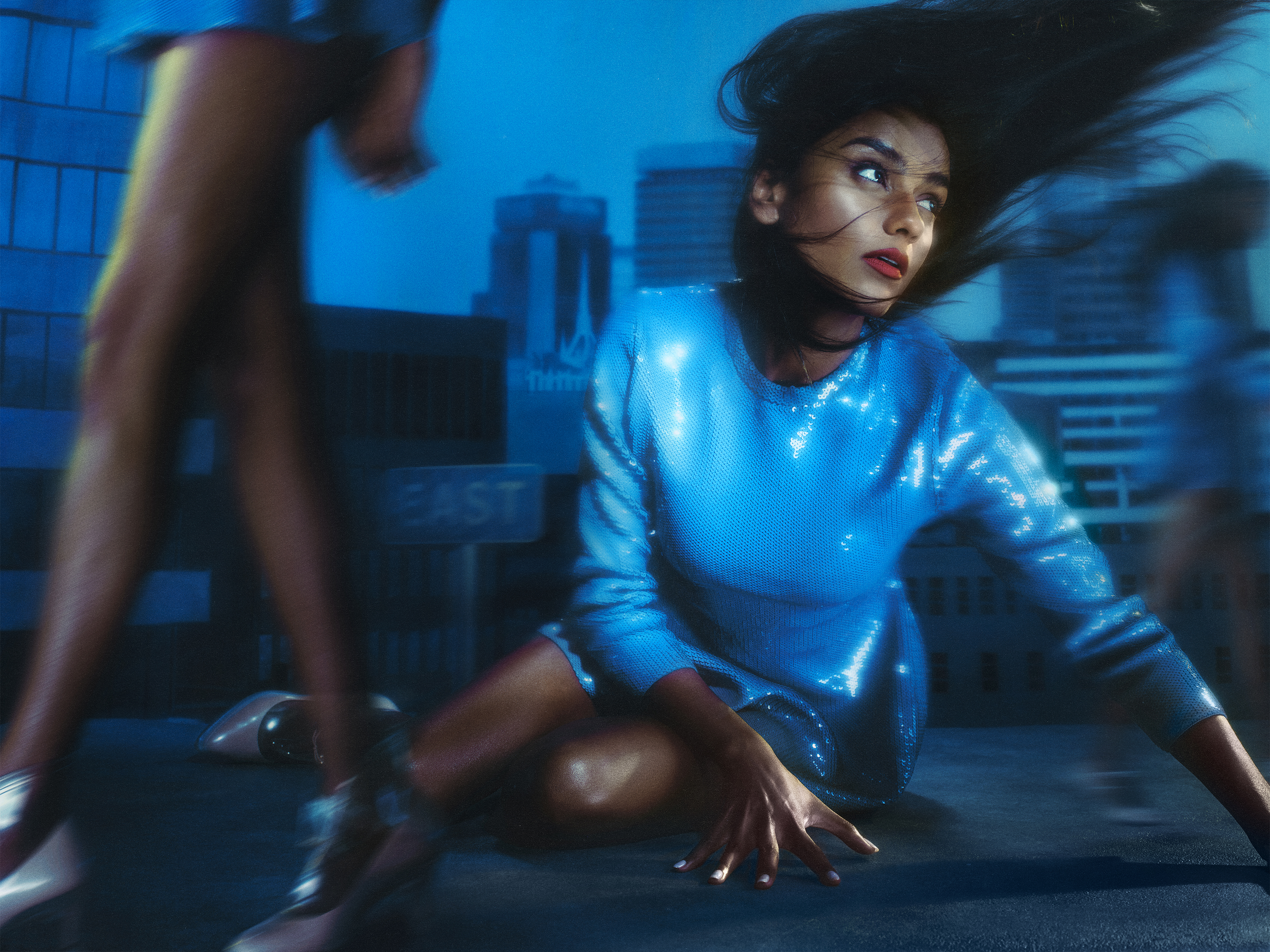 Simone Ashley sitting in front of a cityscape backdrop wearing a blue sequin midi dress. Her hair is being blown to the side.