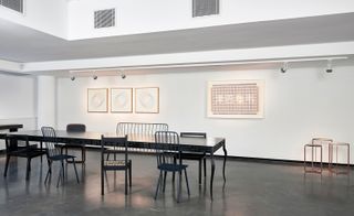 View of a space with white walls, grey floors, white bar spotlights, wall art, three side tables and a long dark coloured table with chairs in different styles