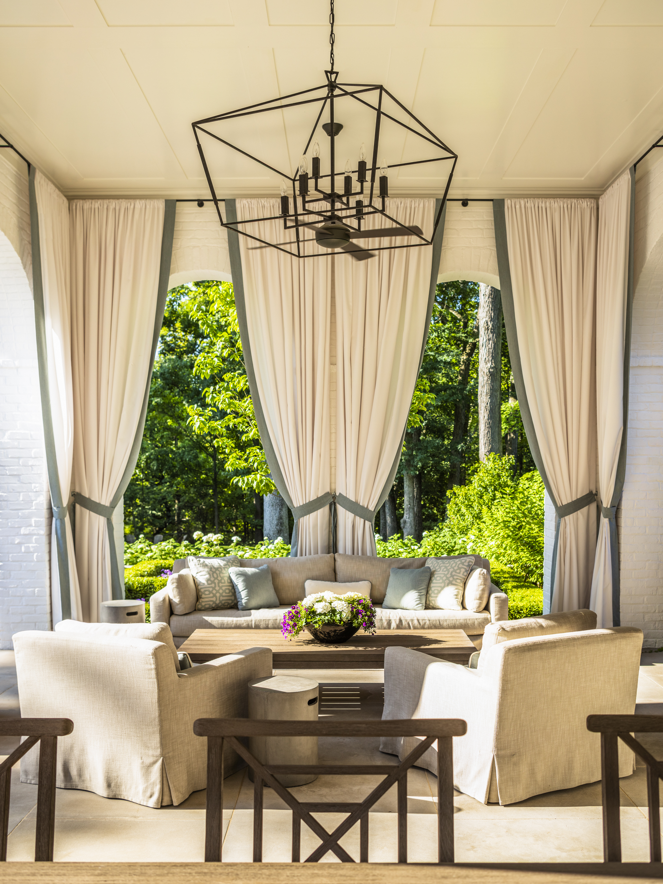 A pergola area with an outdoor sofa, two chairs, and curtains with sage trim hanging up
