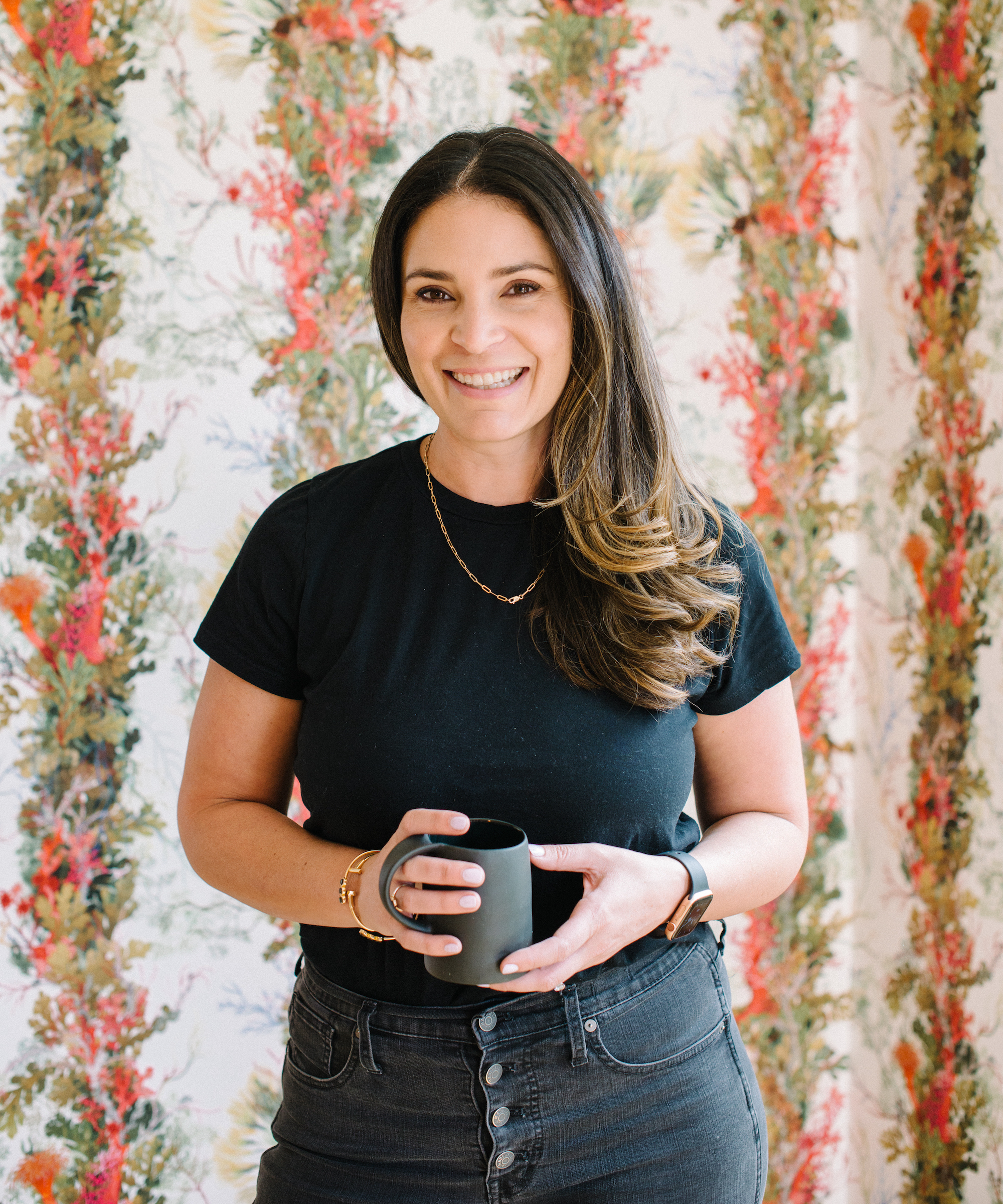 Elsa Ebert, founder and CEO of Composed Living is a woman with long dark hari in a black T-shirt and jeans standing in front of a floral trail wallpaper