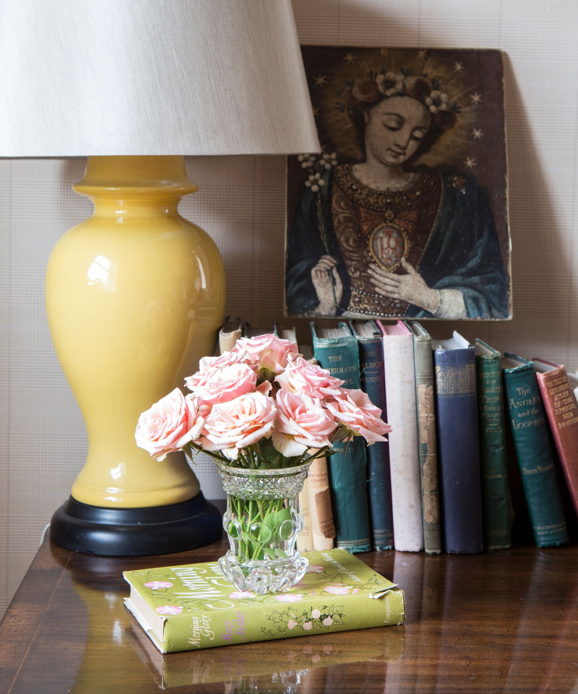 Books on a shiny wooden table with roses, a yellow ceramic lamp with white lampshade, a glass vase of pink roses