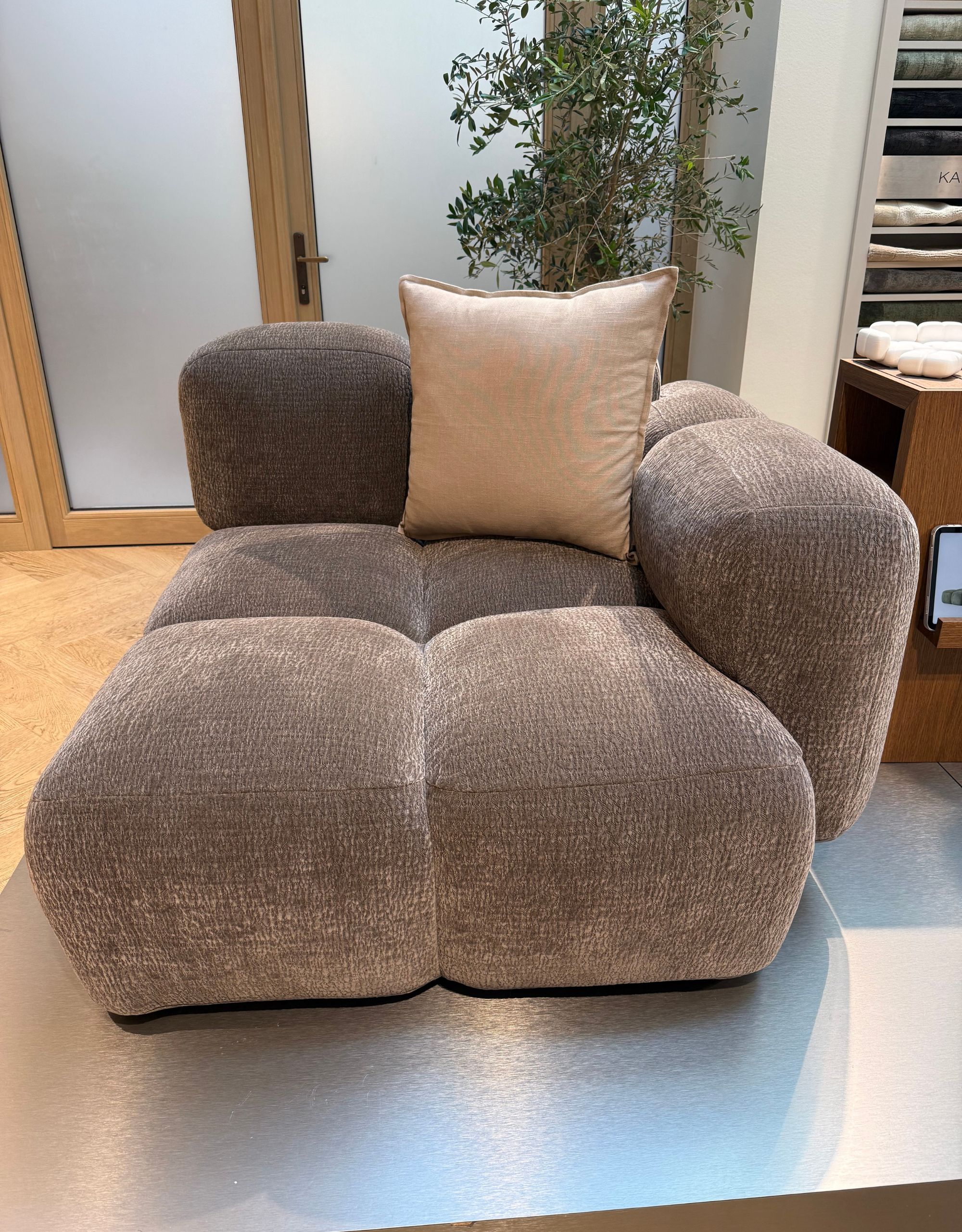 A corner module of a bubble-style contemporary sofa in a pale brown color with a beige cushion in the corner in front of closed double doors and a potted plant