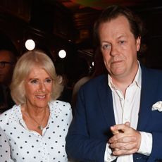 Queen Camilla wearing a white polka dot dress smiling and posing with her son Tom Parker Bowles who is wearing a nvay suit and white shirt