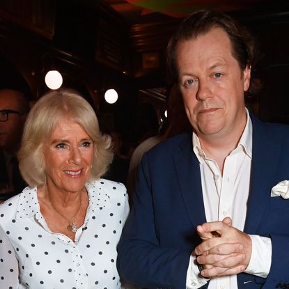 Queen Camilla wearing a white polka dot dress smiling and posing with her son Tom Parker Bowles who is wearing a nvay suit and white shirt