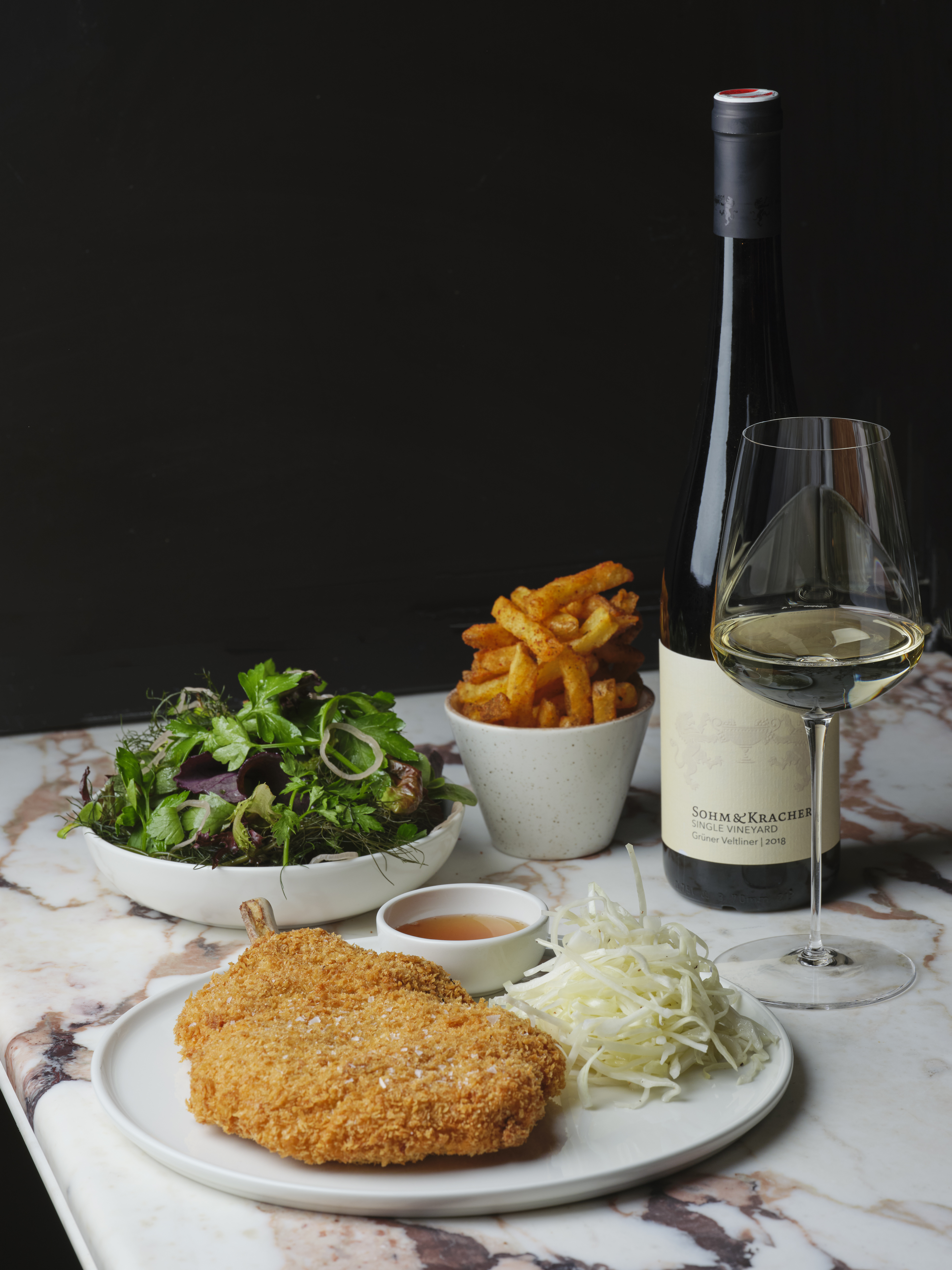 A schintzel served with white radishes and a red dressing, salad, and a portion of paprika-topped fries sits next to a white wine glass and a bottle on a purple marble-topped table.