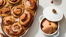 A close up of some fresh cinnamon rolls in a heart shaped pan, next to a small white ceramic pot filled with cookies, with the lid partially off. The lid has a metal heart shaped handle. 
