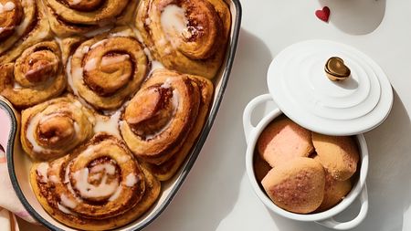 A close up of some fresh cinnamon rolls in a heart shaped pan, next to a small white ceramic pot filled with cookies, with the lid partially off. The lid has a metal heart shaped handle. 
