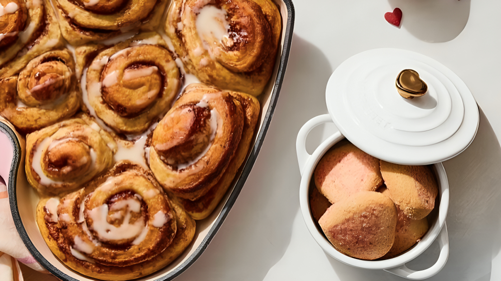 A close up of some fresh cinnamon rolls in a heart shaped pan, next to a small white ceramic pot filled with cookies, with the lid partially off. The lid has a metal heart shaped handle. 