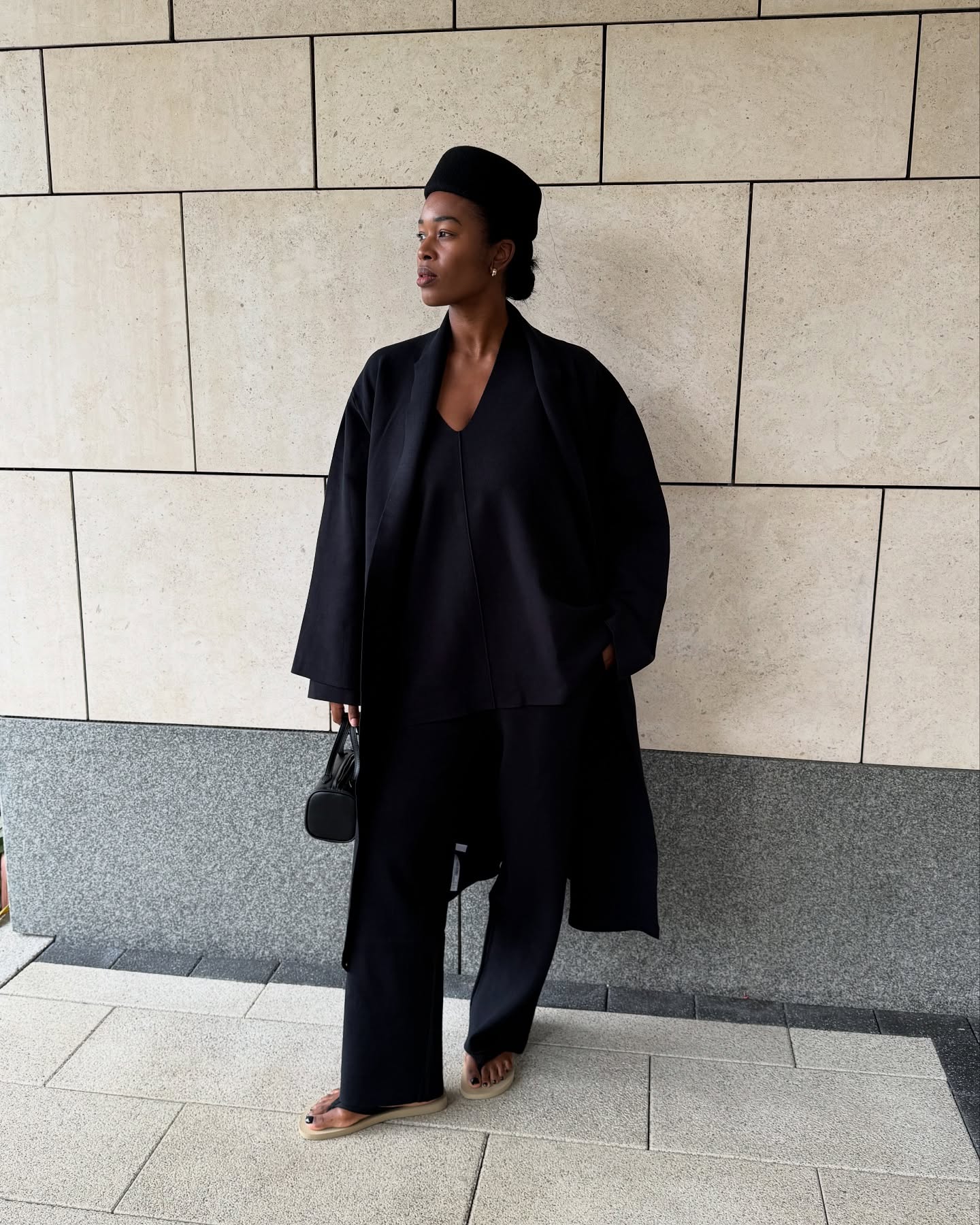 An image of @nlmarilyn wearing a pillbox hat with a black coat and The Row&#039;s dune flip flops.