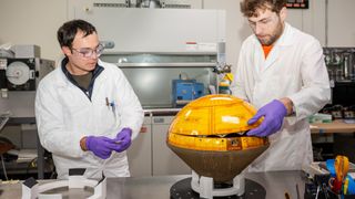 two men in white lab coats handle a yellow cone-shaped capsule on a metal workbench