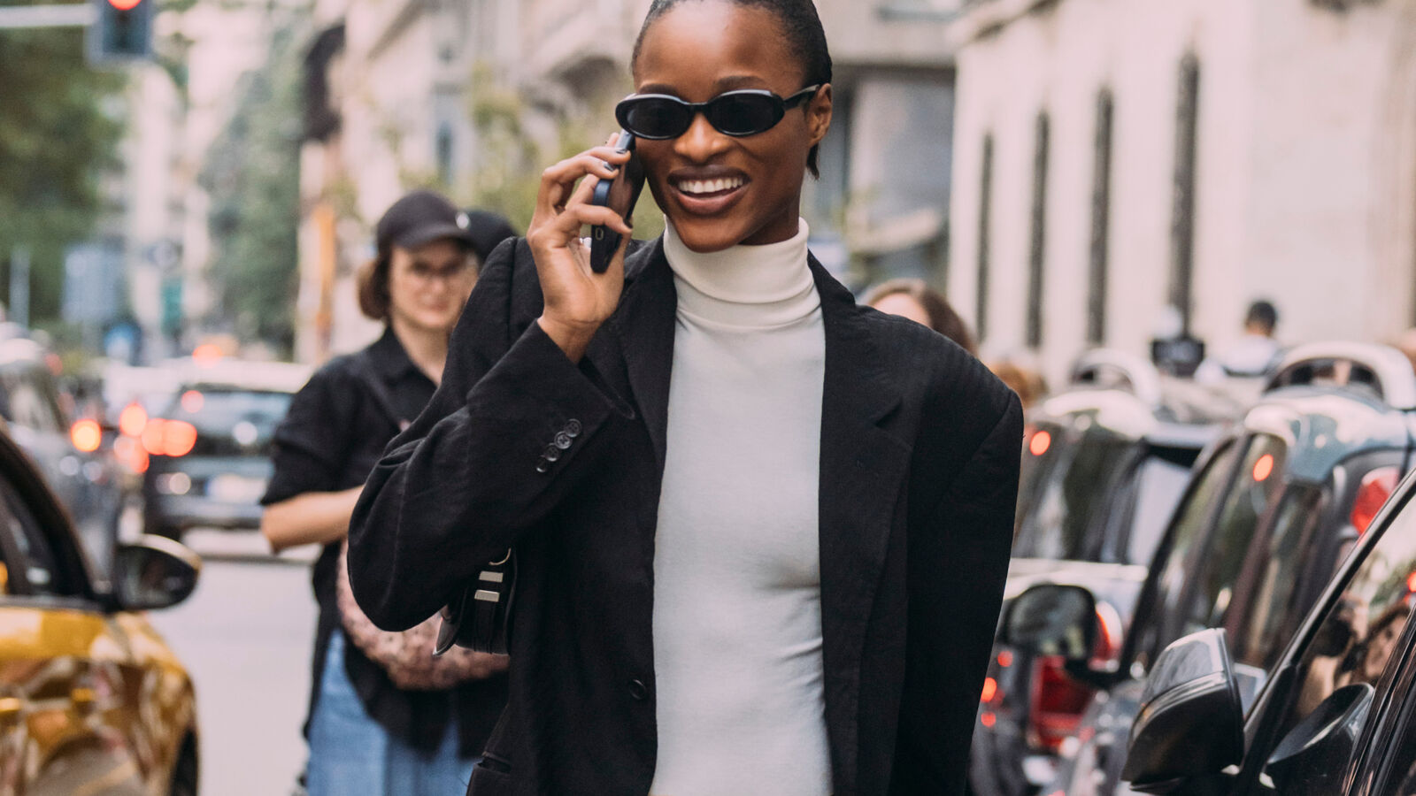 Woman on the phone wearing a white turtleneck, black blazer, and sunglasses 