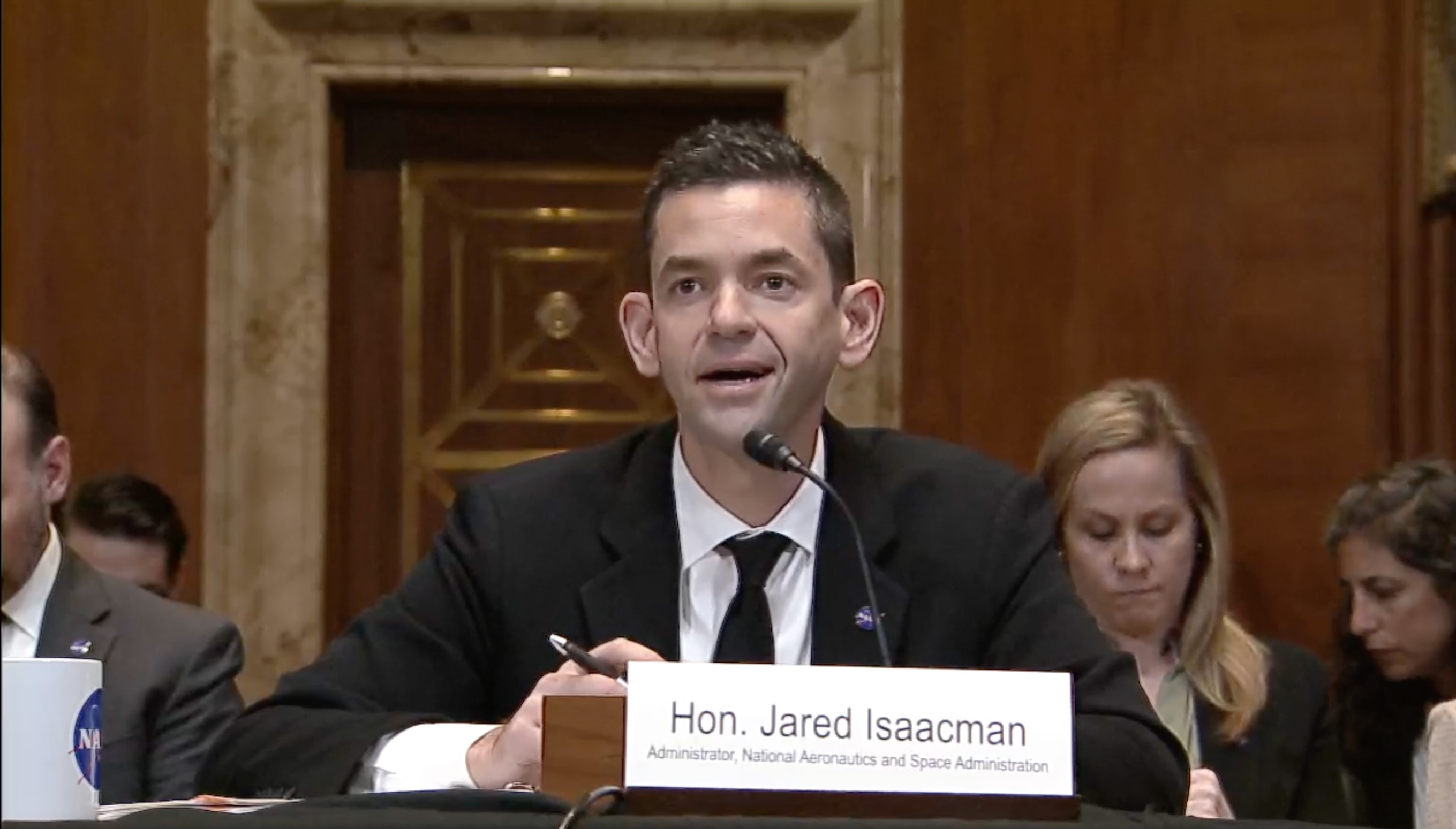a man with big ears wearing a suit speaks into a microphone while sitting at a table in a fancy conference room