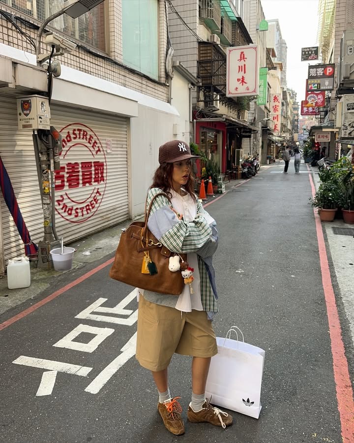 A woman standing wearing baggy shorts, a flannel, a baseball cap, and sneakers.