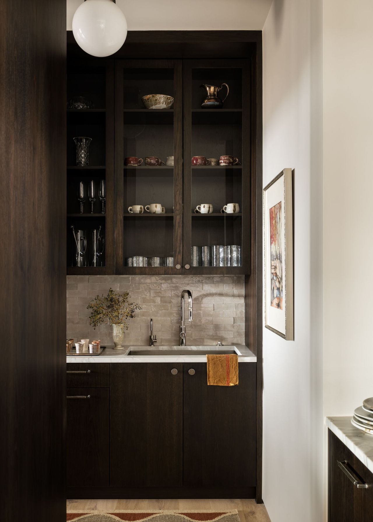 A butler's pantry in dark wood tones with glass-fronted cabinets with a rug on the floor and decorative items on the shelves