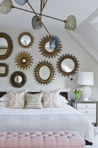 White bedroom with gallery wall of mirrors, metal and white bedside, white table lamp with textured base, pastel patterned cushions, pastel pink button backed ottoman