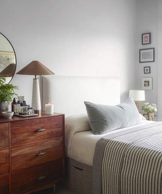A gray bedroom with a brown chest of drawers with a lamp on and a white bed with gray bedding