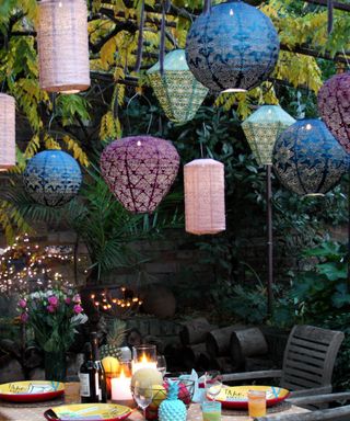 A pergola with pink, green, and blue lanterns hanging from it, plants around it, and a wooden dining table with colorful plates, candles, and bottles of wine on it