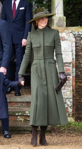 Kate Middleton wearing a long green coat and brown boots with a green hat holding hands with Prince Louis outside church