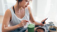 Woman sipping green smoothie and eating bowl of nuts, looking at phone to find out how long will it take to lose a stone in a healthy way