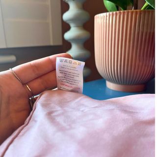 close up of a salmon pink pillow case care label with a ribbed terracotta plant pop in background and a bobby lamp in white visible