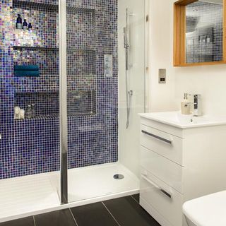 bathroom with black floor tiles, a large walk in shower with blue mozaic tiles on the wall and a square mirror with wooden frame above a white vanity unit