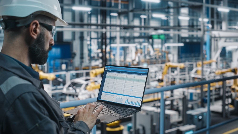A man wearing a helmet and protective glasses overlooks industrial operations and enters information onto a laptop.