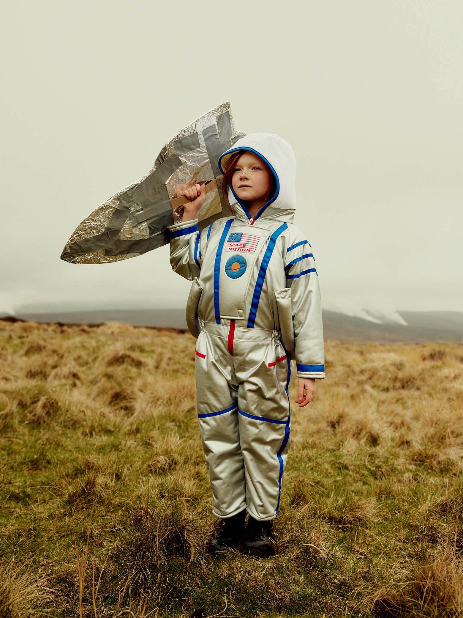 A child in a shiny astronaut costume stands on a grassy field, holding a silver, homemade spaceship. The sky is overcast, creating a dreamy, imaginative atmosphere