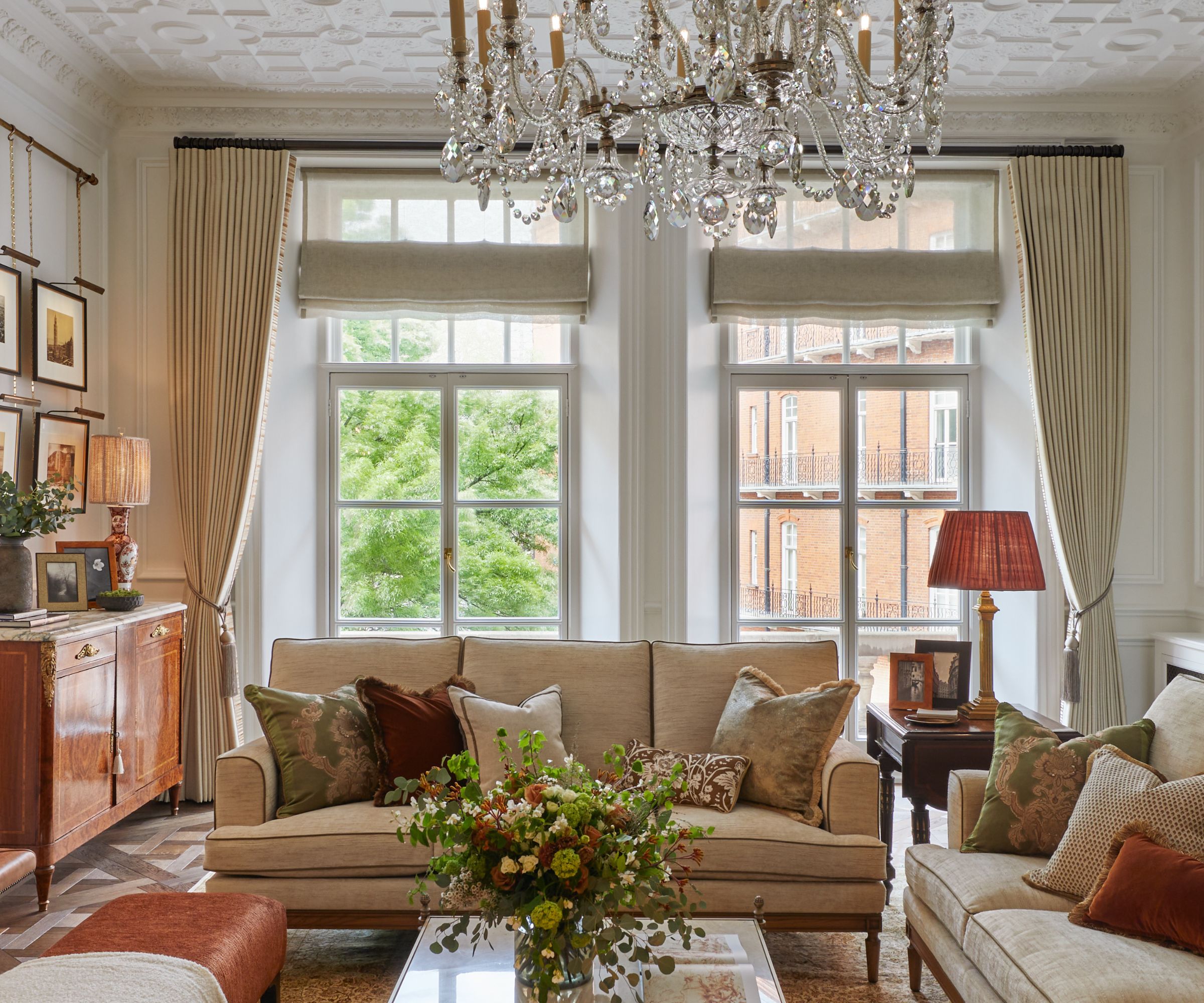 Traditional-style living room with large windows, beige sofa with cushions, coffee table with vase of flowers in front, side tables with lamps to the side
