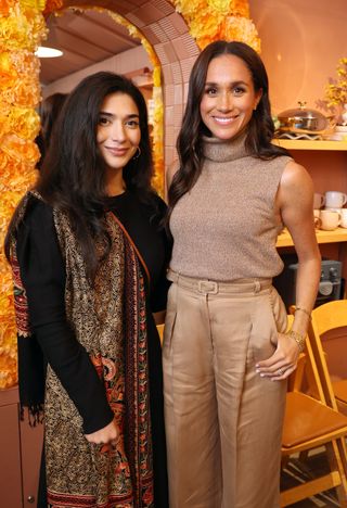 Meghan Markle wearing tan pants and a matching sleeveless turtleneck posing in front of shelves of cookware with Our Place owner Shiza Shahid, wearing a black top and shawl