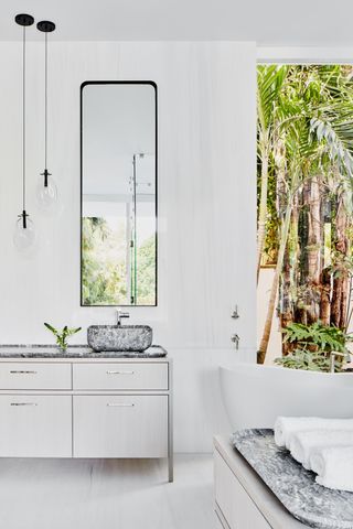 A bathroom with white walls and elements, and large windows that showcase the leafy outdoors