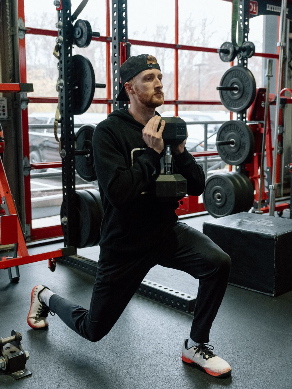 man in black hoodie and trousers performing a reverse lunge holding a dumbbell at his chest in a gym setting with a row of red squat racks behind him. 