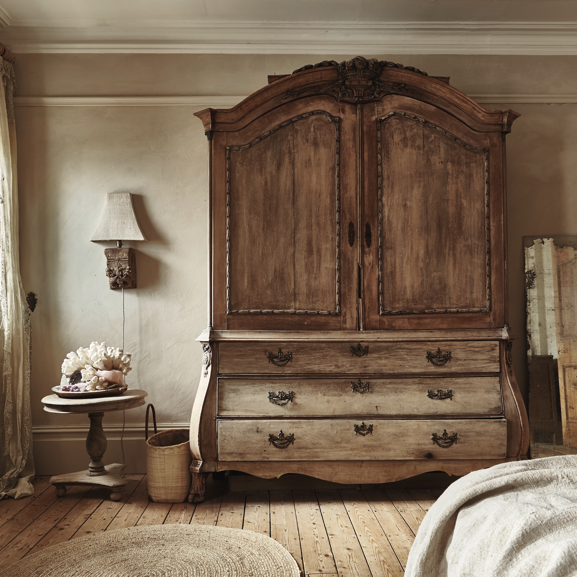 neutral bedroom with bare wooden floor with rug, large vintage armoire and circular side table with a display of coral