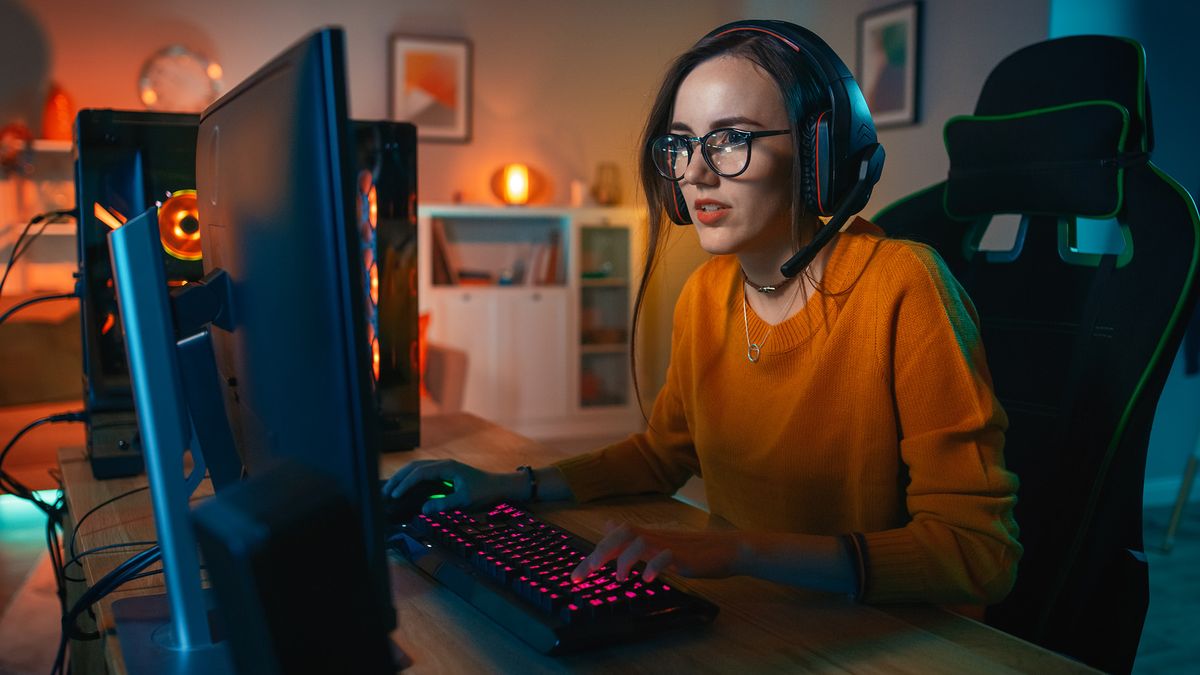 A woman wearing an orange top sitting at a desktop computer with a headset on playing video games