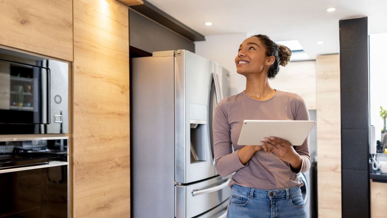 A smiling woman uses a table in her kitchen and looks toward the ceiling.