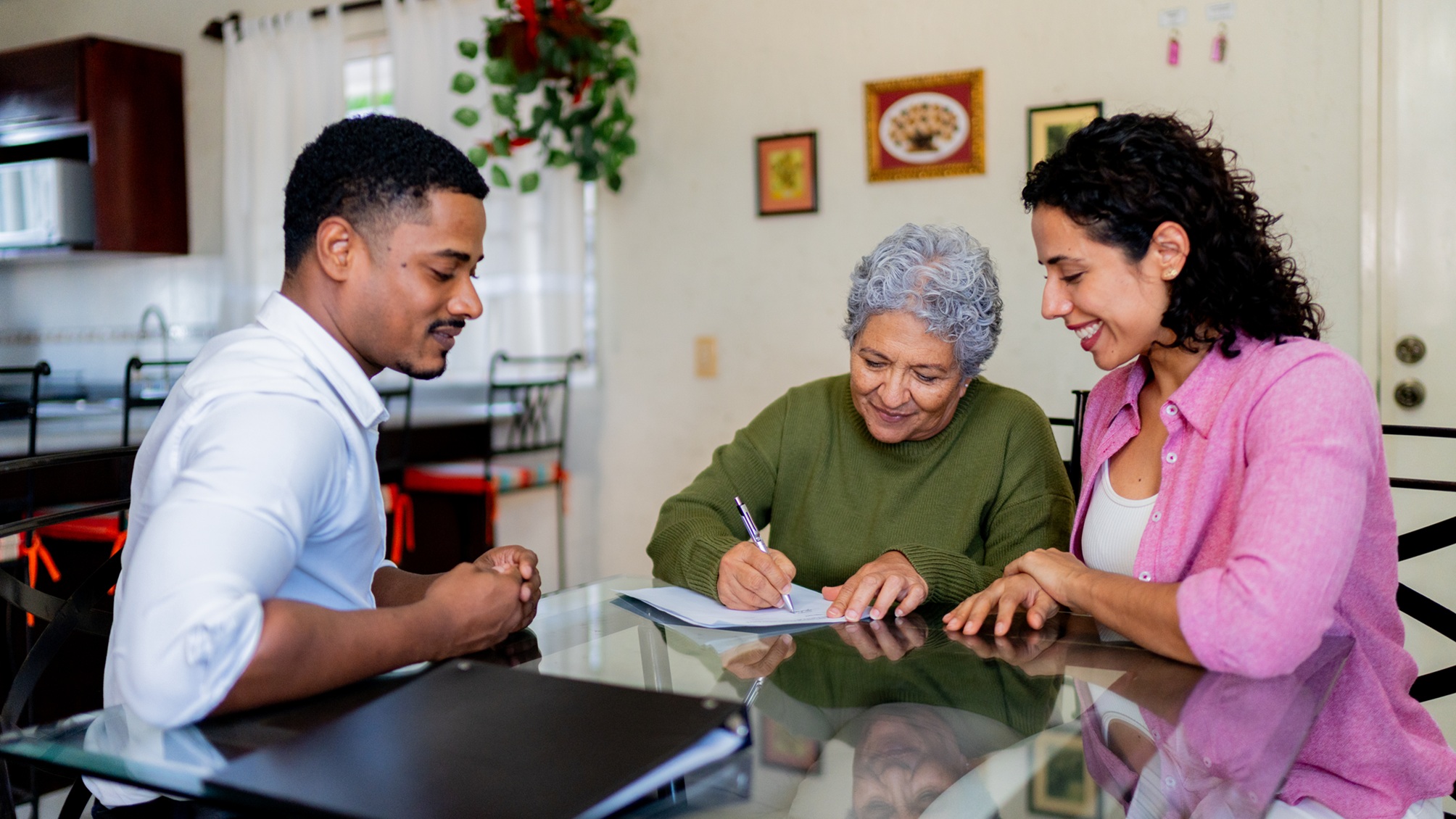 A financial adviser watches as an older woman signs paperwork while her adult daughter looks on at the kitchen table.