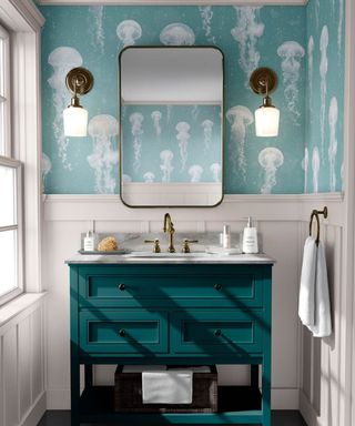 A small bathroom with light blue jellyfish wallpaper, a rectangular mirror, two wall sconces, white shiplap paneling, and a teal blue sink with a gray marble countertop and gold faucet