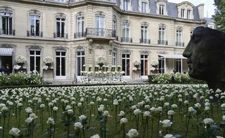 Daytime outside image of Paris' Hotel George V, lawned filled with white roses, white stone building, multiple windows, black framed balconies under first level windows, large male head stone statue to the front right, white roses decorate the front of the building