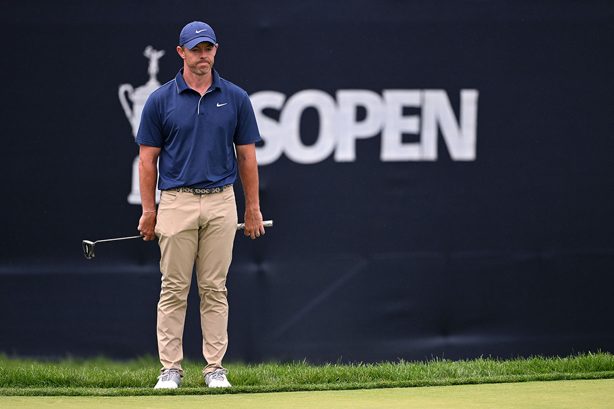 Rory McIlroy in front of a US Open sign, holding his putter behind his back and looking disappointed or frustrated