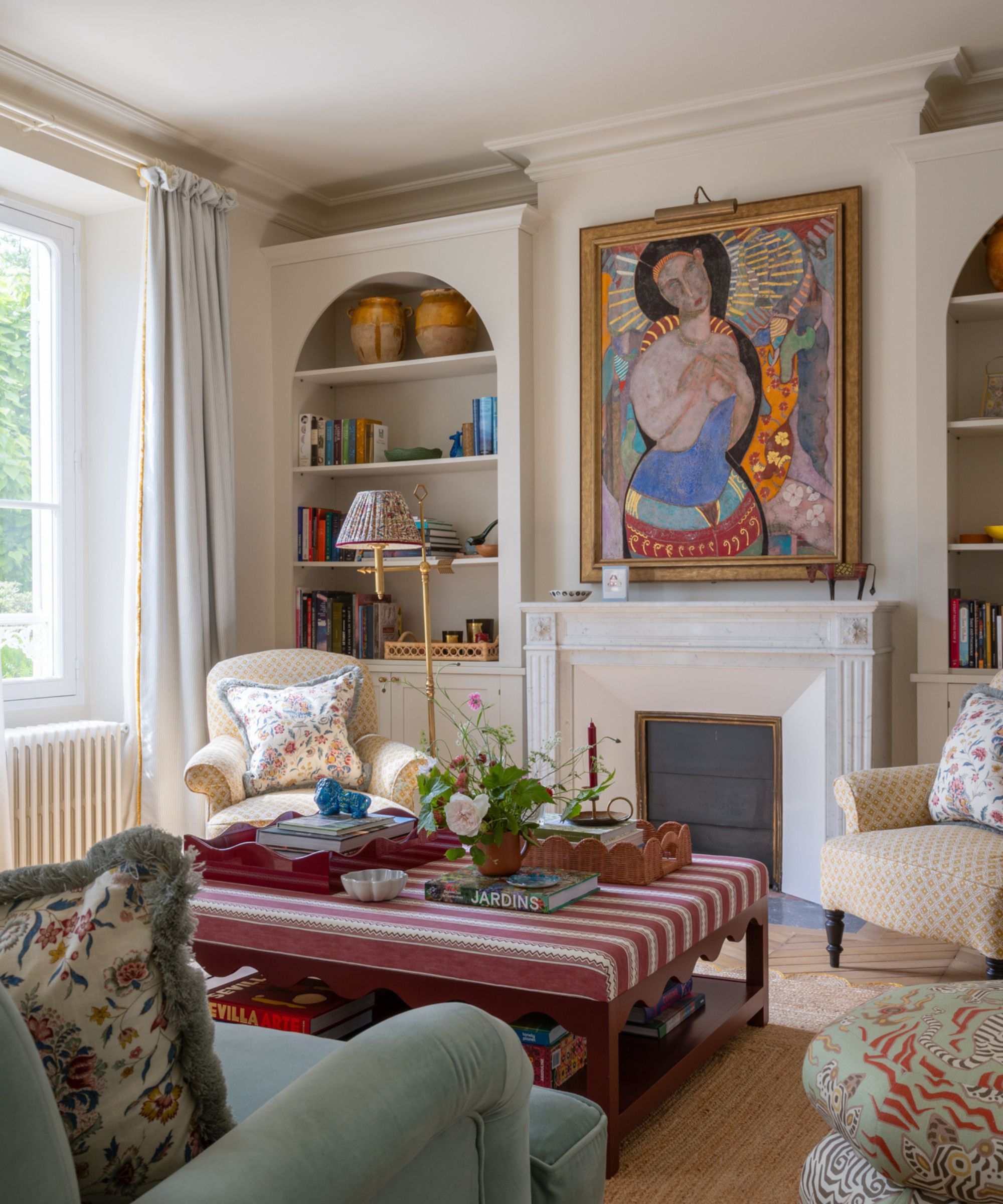 A small living room with cream walls and a white fireplace, flanked by built-in shelves, a large colorful painting above the mantel, floral armchairs, and a red striped coffee table.