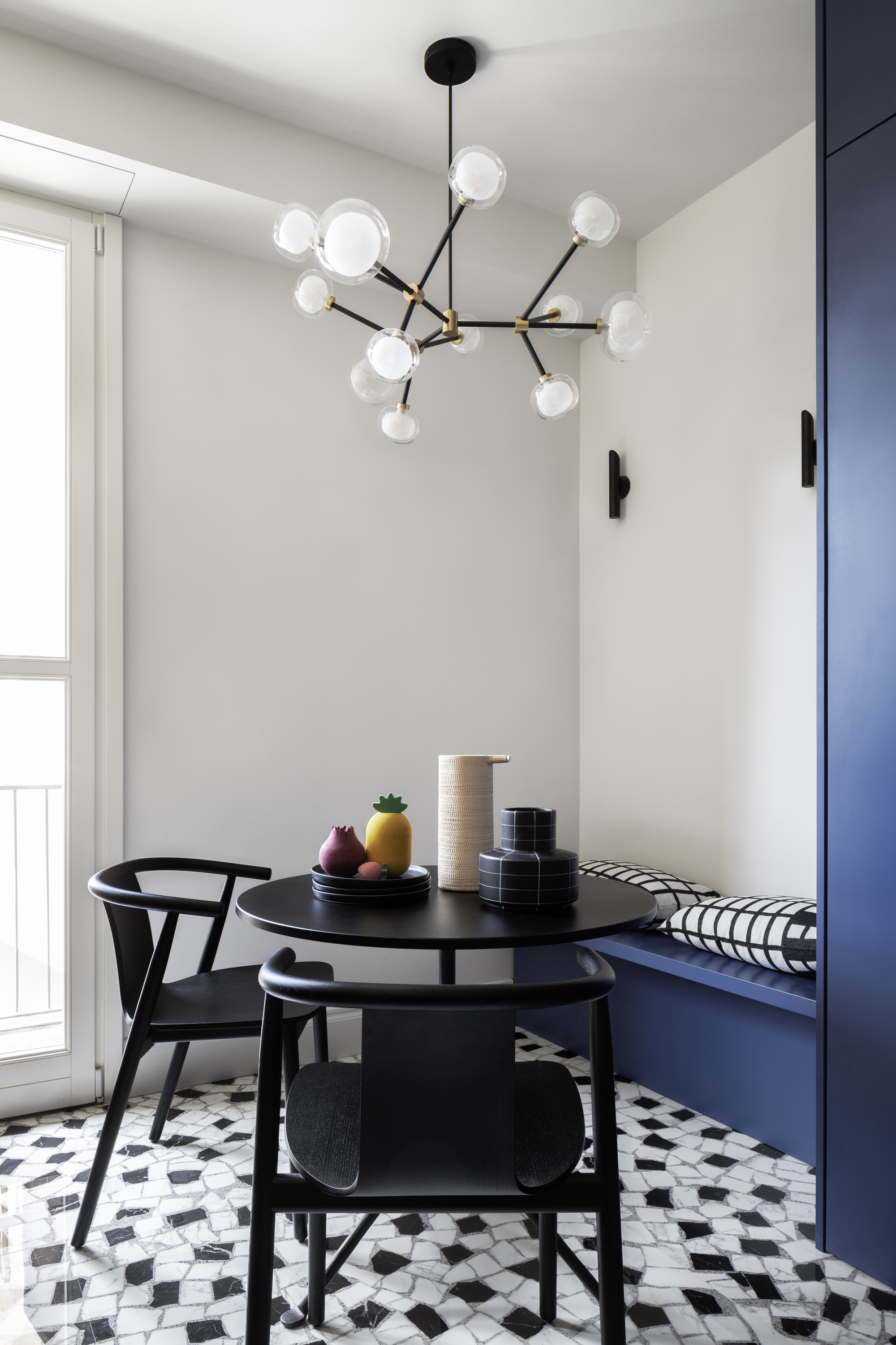 Image of a dining area in a white kitchen. The table is a small, round black table with two matching chairs and a bright blue bench seat. There is a pendant light over the table, and the flooring is made of black and white palldiana terrazzo tile.