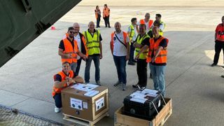 a dozen people stand on an airport tarmac behind two small cargo pallets