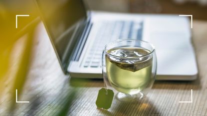 A cup of fresh green tea in a glass mug next to a laptop on a desk as one of the healthy alternatives to coffee
