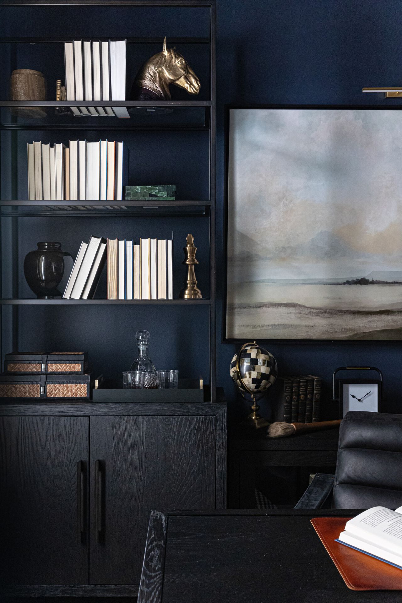 Image of a navy blue room that has a slim, black shelving and storage unit full of books and trinkets. There is a black desk and chair in view and a light landscape painting next to the shelf.
