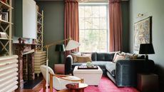 A living room with teal walls, a bright pink rug, and a teal velvet corner sofa in front of a large window.