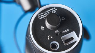 a black microphone by cherry XTRFY with a branded muff and an XLR and USB-C port sits atop a pale brown desk with a dark blue background