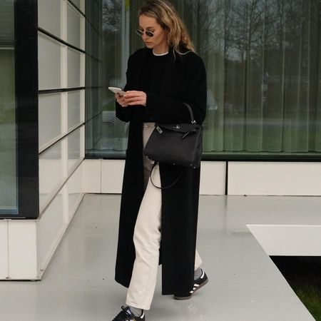 style influencer Anouk Yve wearing wire-rimmed sunglasses, a black coat, black crewneck sweater layered over a white tee, small black Hermes bag, ivory jeans, gray socks, and black Adidas sneakers while looking at her phone