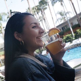 Woman in a denim jacket and hoop earrings drinking a beverage by the pool.