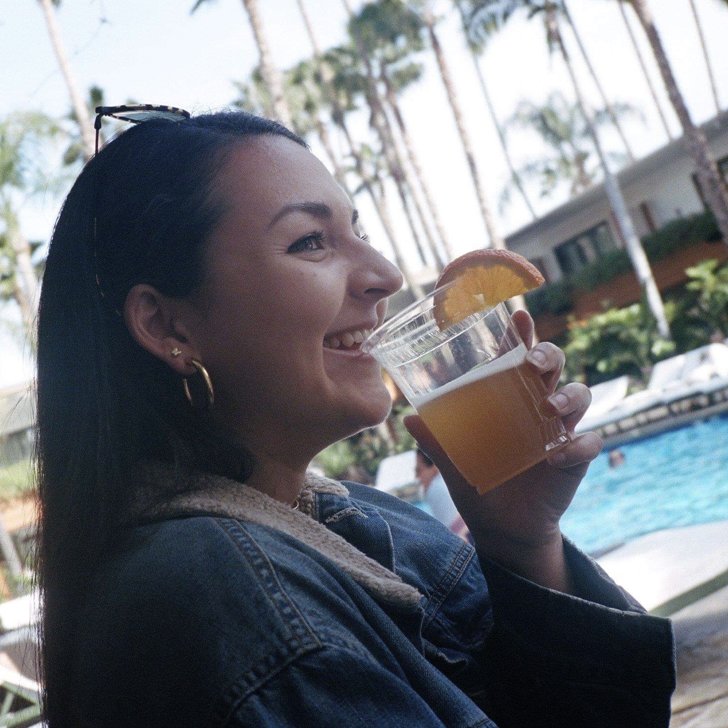 Woman in a denim jacket and hoop earrings drinking a beverage by the pool