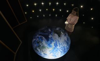 View of a woman holding a small bowl of soup in front of an illuminated model of the earth in a darkly lit space. There are also lights that resemble stars